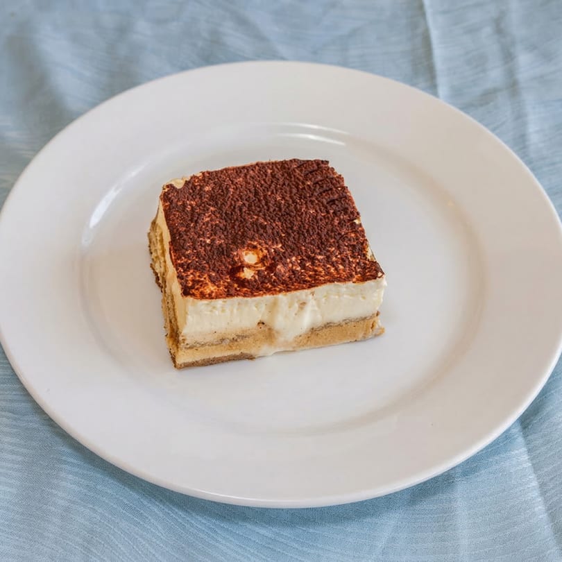 A slice of tiramisu with cocoa powder on top, served on a white plate.
