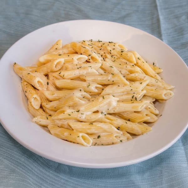 Penne pasta with creamy white sauce and herbs in a white bowl on a blue tablecloth.